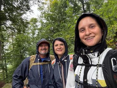 Drei lächelnde Wanderer in Regenjacken mit Kapuzen stehen in einem grünen Wald. Sie tragen Rucksäcke auf der Via Alpina in der Ostschweiz.