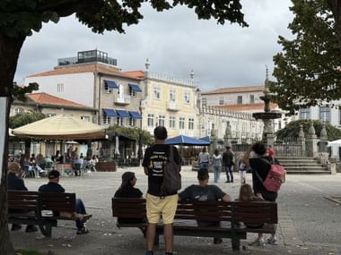 Menschen auf Bänken auf einem Dorfplatz in Caminha, Portugal. Historische Gebäude mit orangen Dächern und ein Brunnen in der Mitte.