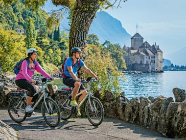 Deux cyclistes roulant le long du rivage du lac Léman avec le Château de Chillon visible sur l'eau. Montagnes et arbres encadrent le chemin.
