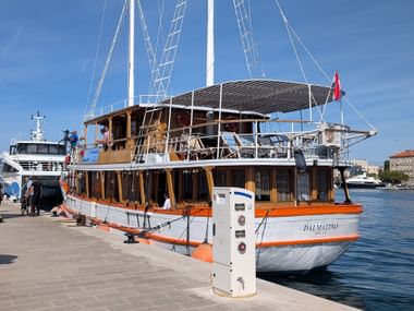 Zweimastiges Segelschiff MS Dalmatino mit weißem Rumpf und orangefarbenen Akzenten am Steinkai unter blauem Himmel in Norddalmatien.
