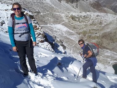 Zwei Wanderinnen stehen bei strahlender Sonne und kurzen Ärmeln im frischen Schnee auf dem Berg.