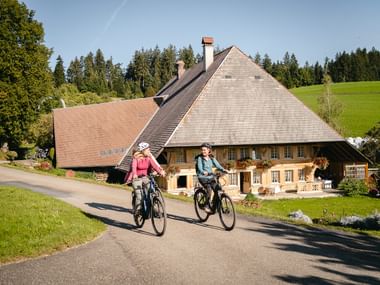 Zwei Velofahrerinnen auf E-Bikes fahren an einem typischen Schweizer Bauernhof mit steilem Dach und Holzbalkonen vorbei, umgeben von Hügeln.