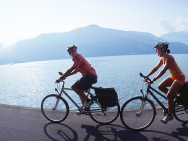 Zwei Radfahrer mit Tourenrädern und Packtaschen fahren entlang einer Seestraße. Berge und ein Segelboot sind über dem Wasser sichtbar.