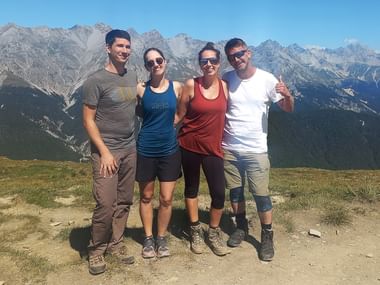 Vier Wanderer stehen auf einem Berggipfel mit felsigen Gipfeln und bewaldeten Tälern hinter ihnen unter klarem blauen Himmel.