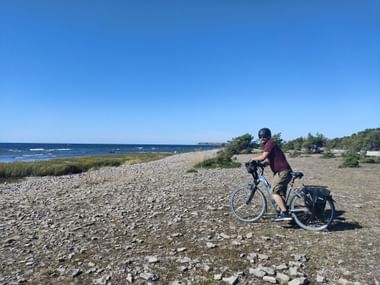 Mann mit Fahrrad steht an einem Kieselstrand mit Ausblick aufs Meer.