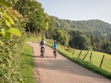 Deux cyclistes roulent sur une route pavée à travers la campagne verdoyante près de Sceut dans le canton du Jura avec des collines boisées.
