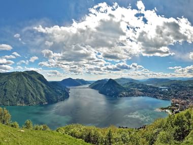 Vue panoramique du lac de Lugano depuis Monte Bré montrant le lac bleu entouré de montagnes vertes sous un ciel nuageux.