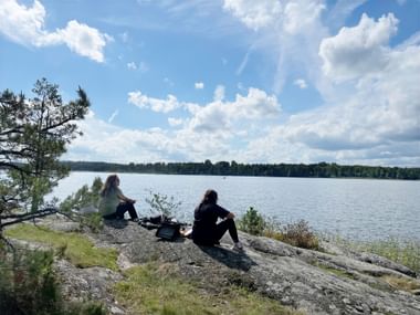 Zwei Frauen sitzen auf felsigem Ufer mit Blick auf einen See mit bewaldeten Inseln. Kiefern rahmen die Szene unter blauem Himmel mit weißen Wolken.