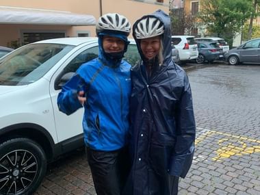 Zwei Frauen posieren vor einem Auto in Regenjacken und Fahrradhelmen.