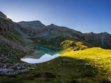 Alpensee in Schweizer Berglandschaft Türkisfarbener Alpensee umgeben von felsigen Bergen und grünen Wiesen im goldenen Morgenlicht mit klarem blauen Himmel.