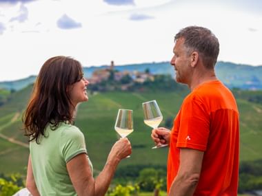 Couple trinquant avec des verres de vin blanc surplombant les collines viticoles et village perché des Langhe du Piémont, Italie.