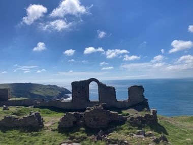 Steinruinen mit Bogenfenster auf Klippe in Cornwall, mit Blick auf blaues Meer und grüne Küstenhügel unter klarem Himmel.