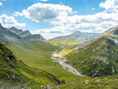 Panoramablick auf die Greina-Ebene mit gewundenem Fluss durch grünes Tal, umgeben von Bergen unter bewölktem blauen Himmel.