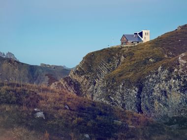 Berghütte auf felsigem Grat im Wallis mit gezackten Gipfeln im Hintergrund unter klarem blauen Himmel.