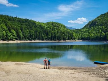 Deux randonneurs avec sacs à dos sur la plage de sable du Nussensee, entourés de collines boisées sous un ciel bleu avec nuages blancs.