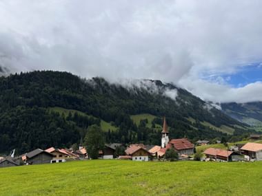 Alpendorf Jaun mit weißer Kirche und rotem Turm in grüner Wiese. Bewaldete Berge mit tiefen Wolken im Hintergrund unter bewölktem Himmel.