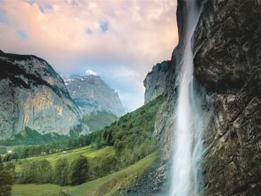 Staubbachfall Wasserfall stürzt eine hohe Klippe im Lauterbrunnental hinab mit grünen Wiesen und dramatischen Berggipfeln unter bewölktem Himmel.
