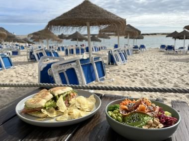Sandwich mit Chips und Poke Bowl mit Lachs auf Holztisch. Strand mit blau gestreiften Liegen und Strohdächern im Hintergrund.