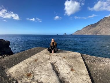 Riana sitzt auf einem Stein und in ihrem Hintergrund liegt die Küste von El Hierro.
