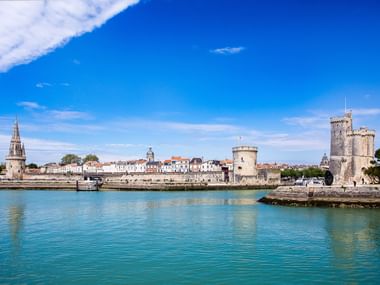 Blick auf den Hafen von La Rochelle mit türkisfarbenem Wasser, historischen Steintürmen und Ufergebäuden unter blauem Himmel.