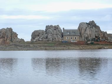 Steinhaus zwischen dramatischen Granitfelsen am Le Gouffre de Plougrescant in der Bretagne, über ruhiges Wasser betrachtet.