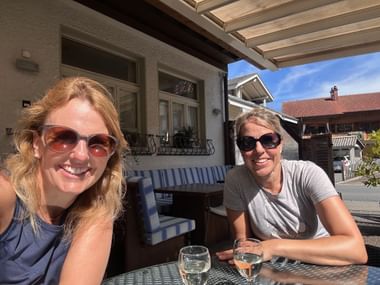 Zwei lächelnde Frauen mit Sonnenbrillen sitzen an einem Tisch im Freien mit Weißweingläsern in Oey, Schweiz.