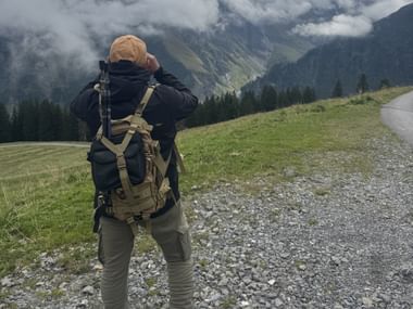 Wanderer mit Rucksack und Mütze auf Schotterweg in Almwiese, fotografiert neblige Berggipfel mit Wäldern in der Ostschweiz.