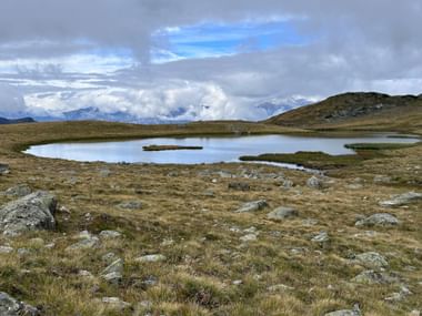 Alpiner Bergsee mit kleinen Inseln, umgeben von felsiger Graslandschaft. Schneebedeckte Berge und dramatische Wolken im Hintergrund.