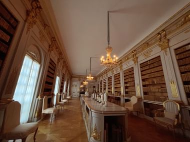 Elegante Bibliothek im Schloss Drottningholm mit raumhohen Bücherregalen, goldenen Verzierungen, Kristalllüstern und antiken Möbeln.