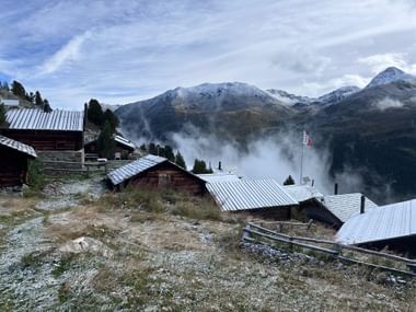 Traditionelle Holzchalets mit Metalldächern in einem Walliser Bergdorf, mit schneebedeckten Alpengipfeln und Nebel im Tal.