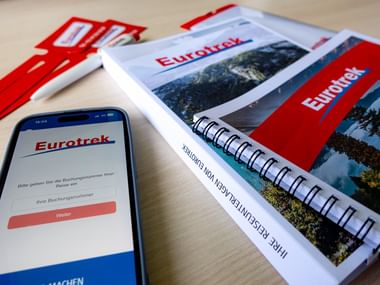 Smartphone mit Eurotrek-Buchungsseite neben spiralgebundenen Reiseunterlagen mit rotem Eurotrek-Branding und Landschaftsfotos auf Holztisch.