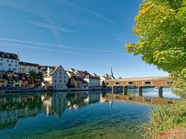 Historische Stadt Diessenhofen am Rhein mit bunten Häusern, Kirchturm und gedeckter Holzbrücke, die sich im ruhigen Wasser spiegelt.