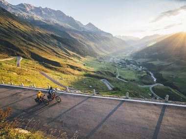 Deux cyclistes gravissent une route de col sinueuse aux Grisons. Vallée alpine avec village en contrebas, pics montagneux et lumière dorée.
