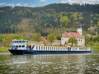 MS Prinzessin Katharina Flusskreuzfahrtschiff fährt auf ruhigem Wasser mit weißer Kirche und bewaldeten Hügeln im Hintergrund unter blauem Himmel.
