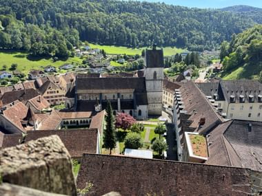 Blick von oben auf die historische Altstadt von Saint-Ursanne mit mittelalterlicher Kirche, traditionellen roten Ziegeldächern und grünen Hügeln.