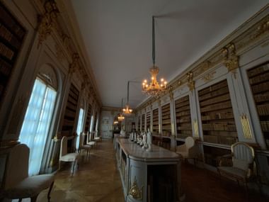 Eleganter Bibliotheksraum im Schloss Drottningholm mit raumhohen Bücherregalen, goldenen Verzierungen, Kristalllüstern und weißen Stühlen.