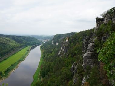 Blick von Sandsteinfelsen auf die Elbe, die sich durch grünes Tal in der Sächsischen Schweiz windet. Bewaldete Hügel und Felsformationen rahmen den Fluss.