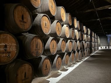 Lange Reihen gestapelter Sandeman-Portweinfässer in einem traditionellen Weinkeller in Porto mit Holzdeckenbalken und gedämpfter Beleuchtung.