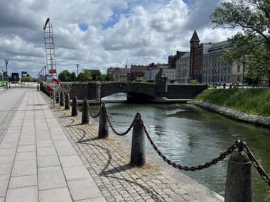 Gepflasterte Uferpromenade in Malmö mit Kettenpoller entlang eines Kanals. Steinbrücke und historische Gebäude mit Turm unter bewölktem Himmel.