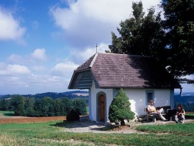 Auf einer grünen Wiese steht eine ganz kleine Kapelle unterb einem Baum. 2 Wanderer sitzen vor der Kapelle und machen eine Pause.