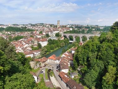 Luftaufnahme von Fribourg mit roten Ziegeldächern, Kathedralturm, Fluss, Steinbrücke und üppigen grünen Bäumen unter bewölktem Himmel.