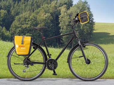 Vélo de location Eurobike noir avec sacoches jaunes et panier avant, garé sur le trottoir avec collines vertes et arbres en arrière-plan.