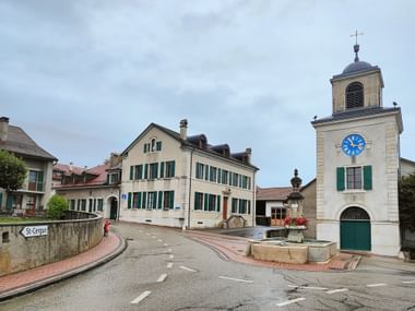 Stadtplatz in Nyon mit weißem Uhrenturm mit blauem Zifferblatt, traditionellen Schweizer Gebäuden mit grünen Fensterläden und Brunnen.