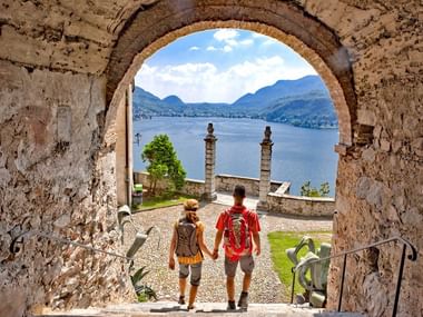 Deux touristes marchant sous une arche de pierre à Morcote surplombant le lac de Lugano avec montagnes et ciel bleu, Tessin, Suisse.