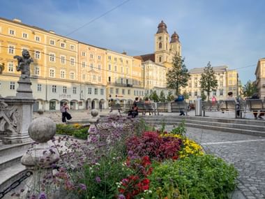 Linzer Hauptplatz mit bunten Blumenbeeten im Vordergrund, gelben historischen Gebäuden und Kirche mit zwei Türmen unter blauem Himmel.