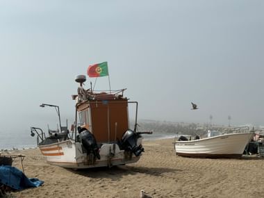 Fischerboot mit portugiesischer Flagge und zwei Außenbordmotoren am Sandstrand bei Vila do Conde. Ein weiteres Boot und Möwe im nebligen Hintergrund.