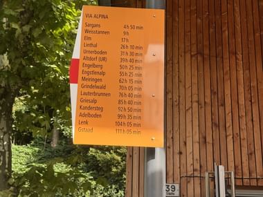 Oranges Via Alpina Wanderschild mit Zielen und Gehzeiten von Vaduz zu verschiedenen Schweizer Orten, an einem Mast befestigt.