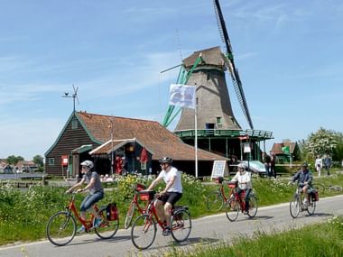 Groupe de cyclistes sur des vélos rouges passant devant un moulin néerlandais traditionnel avec garnitures vertes et bâtiment au toit de chaume.