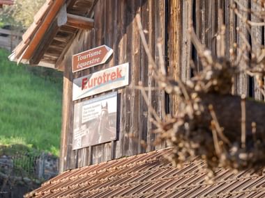 Verwitterte Holzscheune mit Eurotrek-Schild 'Tourisme équestre' an der Wand. Holzschindeldach und verschwommene Äste im Vordergrund.
