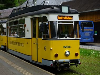 Gelb-weiße Kirnitzschtalbahn Straßenbahn Nummer 6 an einer Haltestelle. Die historische Straßenbahn zeigt ihr Ziel auf dem Frontschild.